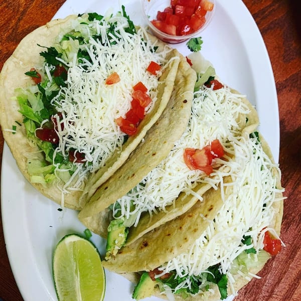 Three Tacos Al Carbon with shredded cheese, lettuce, tomatoes, and a lime wedge on a white plate.