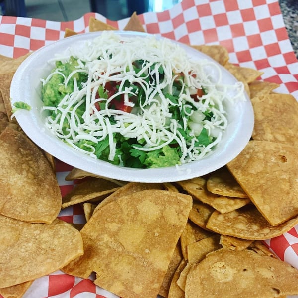 A white bowl of guacamole topped with shredded cheese and pico de gallo, surrounded by tortilla chips.